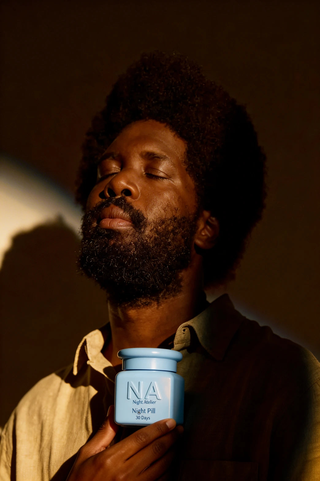 Man holding a blue jar labeled 'NA' by Night Atelier against a dark background
