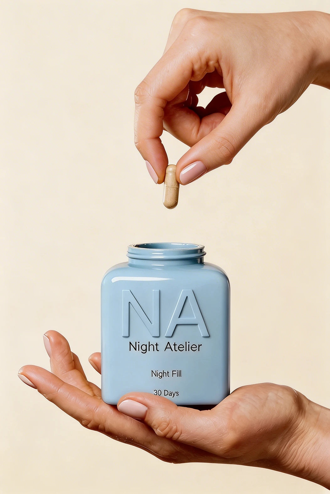 Hand holding a blue container labeled 'Night Atelier' with a pill above it on a beige background