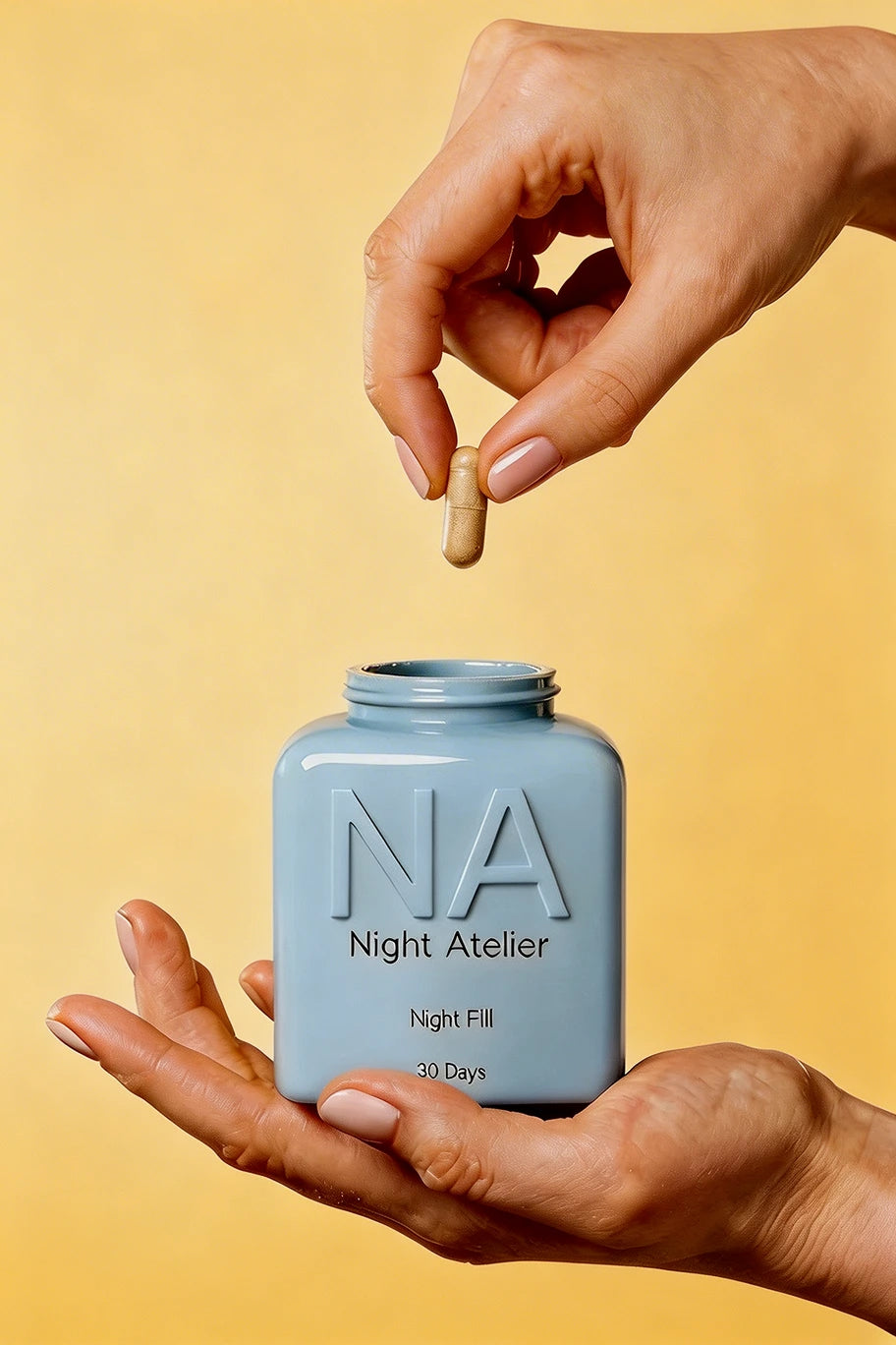Hand holding a small blue container labeled 'NA Night Atelier' with a pill against a yellow background